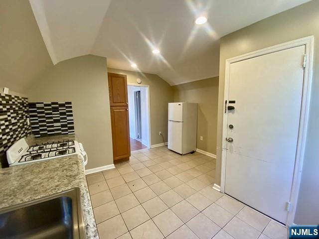 kitchen featuring light tile patterned flooring, white appliances, vaulted ceiling, backsplash, and brown cabinetry