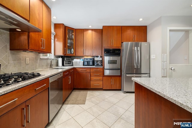 kitchen with light stone counters, a sink, under cabinet range hood, appliances with stainless steel finishes, and a warming drawer