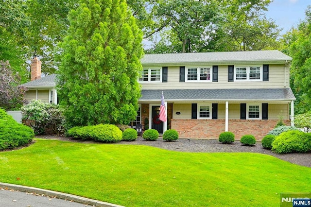 view of front of property featuring roof with shingles, brick siding, a chimney, covered porch, and a front yard