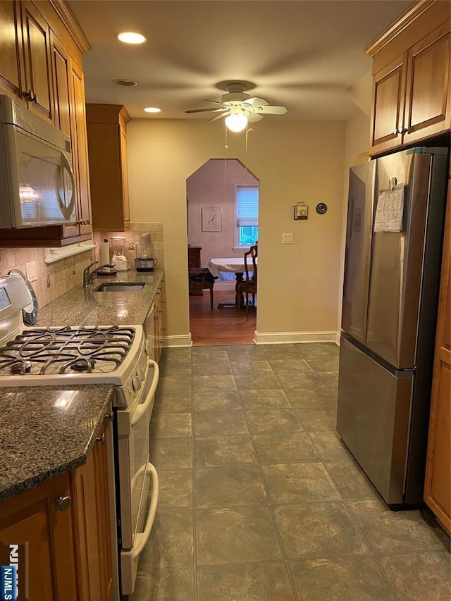 kitchen featuring white appliances, a sink, ceiling fan, backsplash, and dark stone countertops