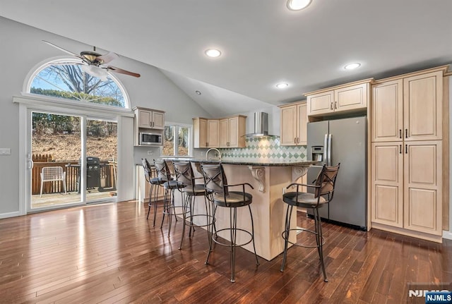 kitchen with a breakfast bar area, dark wood-style floors, appliances with stainless steel finishes, cream cabinets, and wall chimney exhaust hood