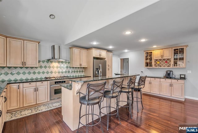 kitchen with dark stone countertops, a breakfast bar, stainless steel appliances, glass insert cabinets, and wall chimney exhaust hood