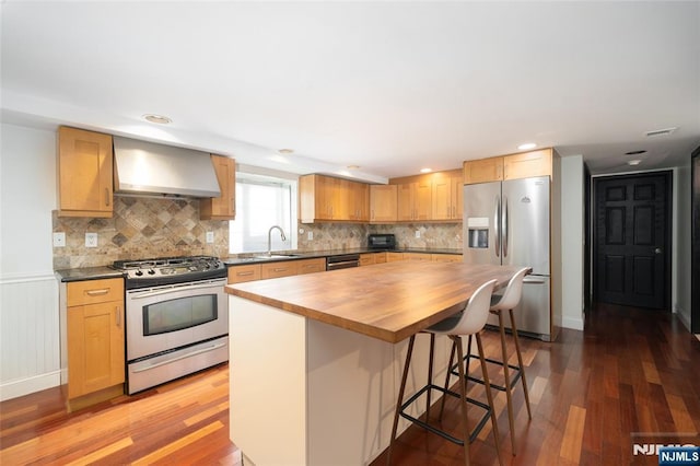 kitchen with stainless steel appliances, wooden counters, wall chimney range hood, wood finished floors, and a kitchen breakfast bar