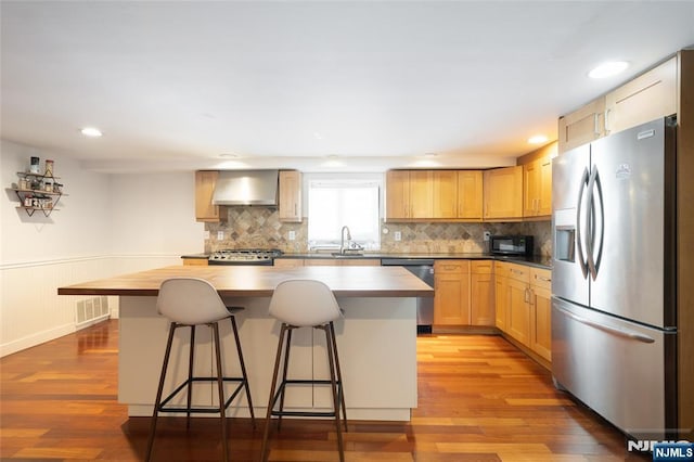 kitchen featuring light wood-type flooring, wall chimney range hood, appliances with stainless steel finishes, and a wainscoted wall