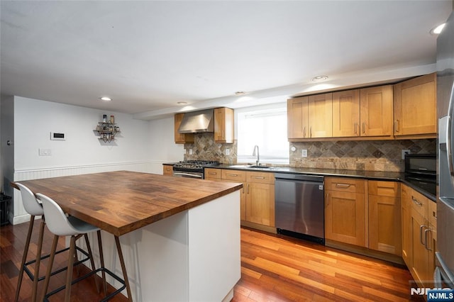 kitchen featuring stainless steel appliances, a sink, wall chimney range hood, butcher block countertops, and light wood-type flooring