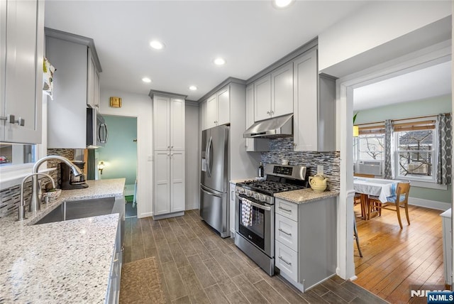 kitchen with gray cabinetry, under cabinet range hood, dark wood finished floors, appliances with stainless steel finishes, and a sink