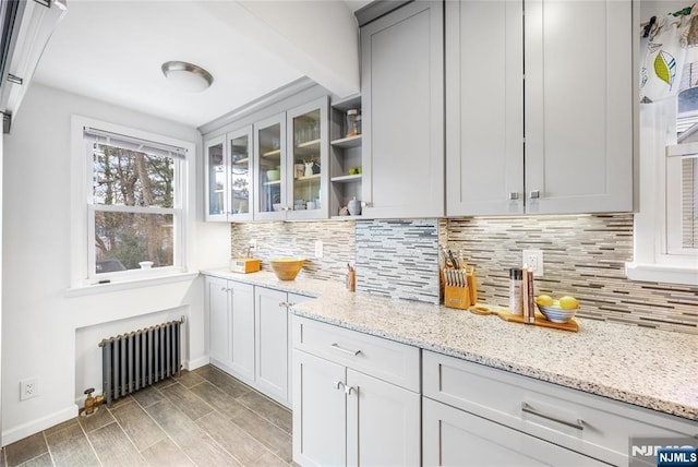 kitchen with glass insert cabinets, radiator heating unit, light stone counters, decorative backsplash, and open shelves