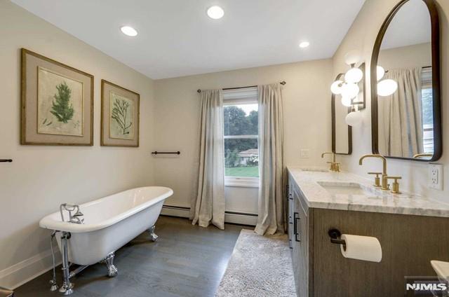 full bath with wood finished floors, double vanity, a freestanding tub, a sink, and baseboard heating