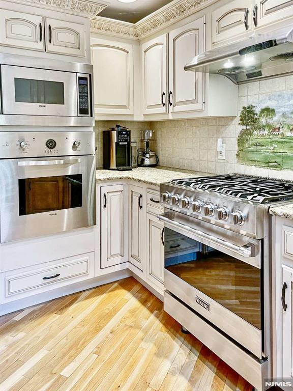 kitchen featuring light wood-style flooring, appliances with stainless steel finishes, light stone counters, under cabinet range hood, and backsplash