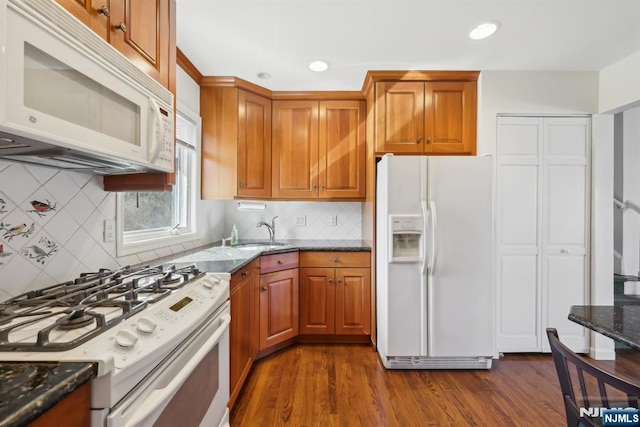 kitchen with dark wood-style flooring, brown cabinetry, a sink, dark stone counters, and white appliances