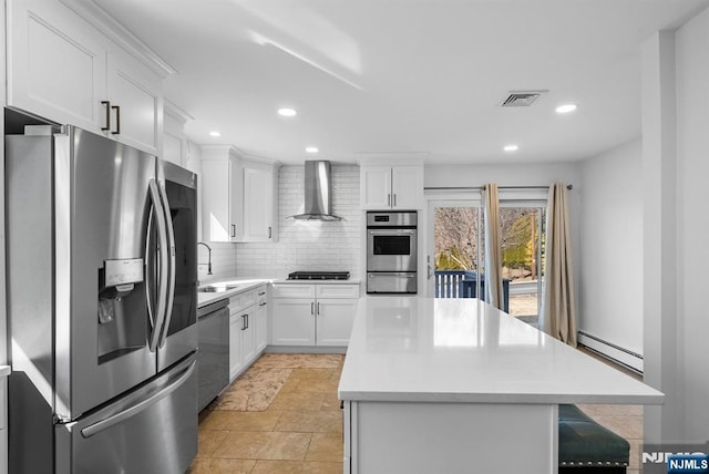 kitchen with baseboard heating, appliances with stainless steel finishes, a sink, a kitchen island, and wall chimney exhaust hood