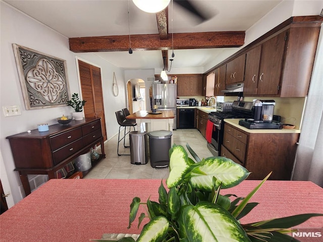 kitchen with arched walkways, under cabinet range hood, light countertops, appliances with stainless steel finishes, and beamed ceiling