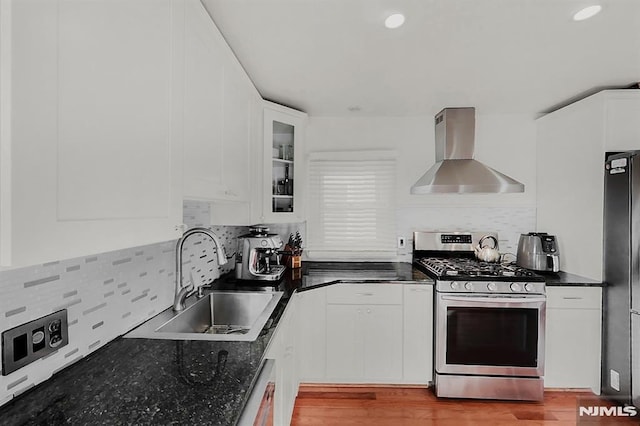 kitchen featuring stainless steel gas stove, freestanding refrigerator, wall chimney range hood, white cabinetry, and a sink