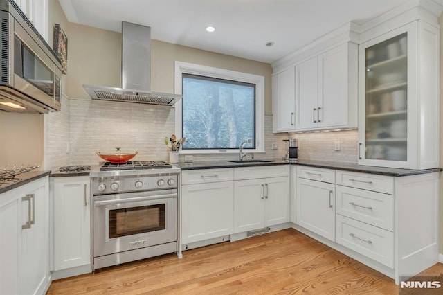 kitchen with light wood finished floors, white cabinets, wall chimney exhaust hood, appliances with stainless steel finishes, and a sink