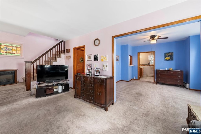 carpeted living room featuring baseboards, a ceiling fan, a glass covered fireplace, stairway, and a baseboard heating unit