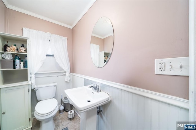 half bath with crown molding, a wainscoted wall, toilet, tile patterned floors, and a sink