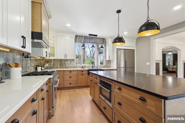 kitchen with arched walkways, stainless steel appliances, decorative backsplash, wall chimney range hood, and brown cabinets