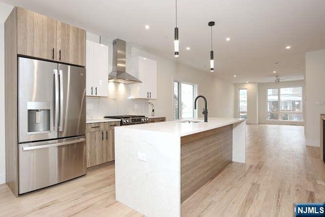 kitchen with a sink, wall chimney range hood, light wood finished floors, stainless steel fridge, and tasteful backsplash