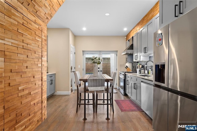 kitchen featuring gray cabinetry, wood finished floors, a sink, appliances with stainless steel finishes, and wall chimney range hood