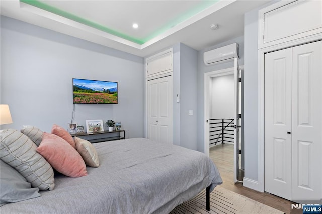 bedroom featuring a raised ceiling, a wall unit AC, light wood-type flooring, two closets, and recessed lighting