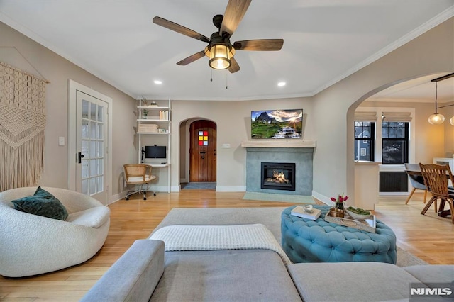 living area with arched walkways, baseboards, ornamental molding, light wood-type flooring, and a glass covered fireplace