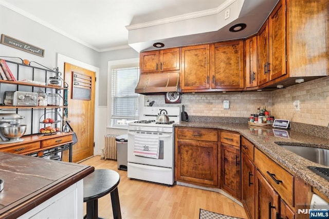 kitchen with radiator heating unit, ornamental molding, brown cabinets, white gas range, and under cabinet range hood