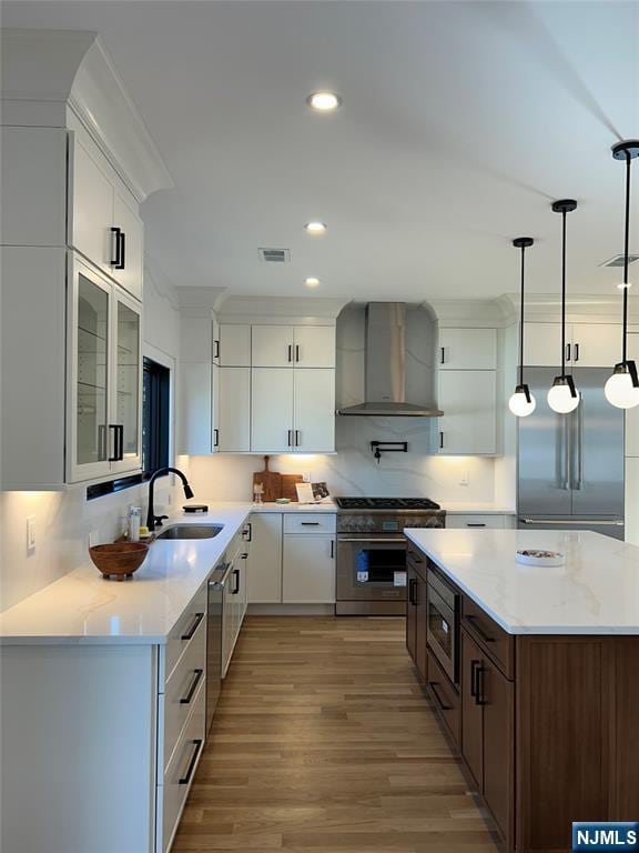 kitchen featuring visible vents, light wood-style flooring, a sink, built in appliances, and wall chimney exhaust hood