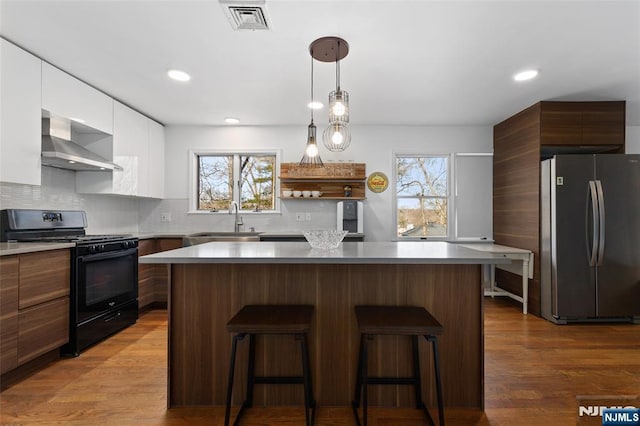 kitchen with black gas range oven, visible vents, wall chimney range hood, freestanding refrigerator, and modern cabinets