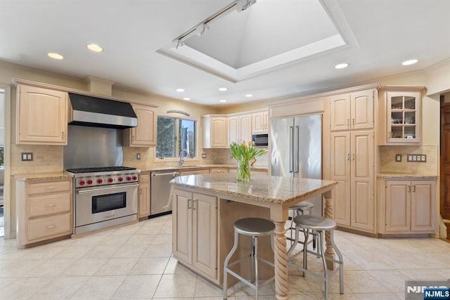 kitchen featuring light brown cabinets, light stone counters, premium appliances, a breakfast bar area, and wall chimney range hood