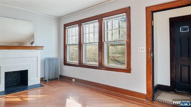 unfurnished living room featuring wood finished floors, baseboards, a brick fireplace, radiator, and crown molding