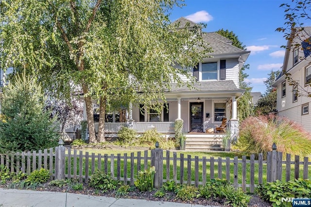 view of property hidden behind natural elements with covered porch, roof with shingles, a fenced front yard, and a front yard