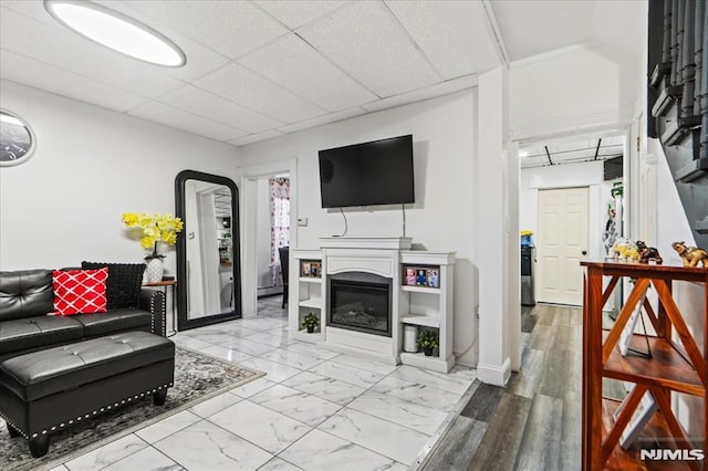 living room with marble finish floor, a drop ceiling, a glass covered fireplace, and baseboards