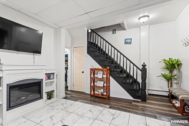 living area with marble finish floor, a paneled ceiling, baseboard heating, a glass covered fireplace, and stairs