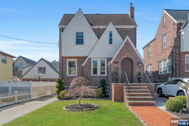 tudor-style house featuring brick siding, fence, roof with shingles, a front yard, and a chimney