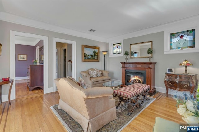 living area with visible vents, crown molding, baseboards, light wood-style flooring, and a glass covered fireplace