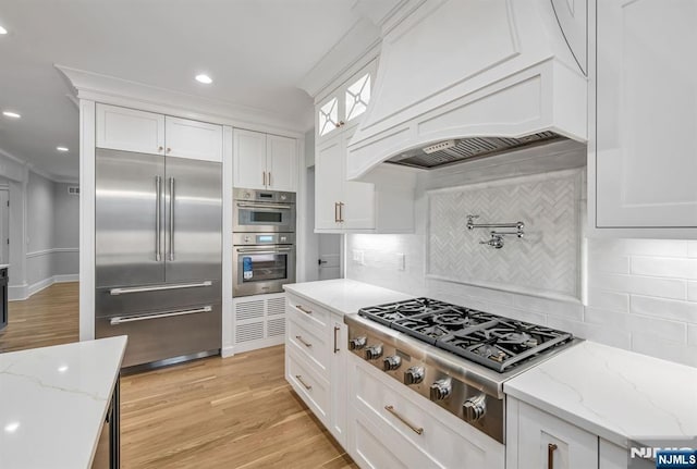 kitchen with premium range hood, white cabinetry, appliances with stainless steel finishes, ornamental molding, and light wood-type flooring