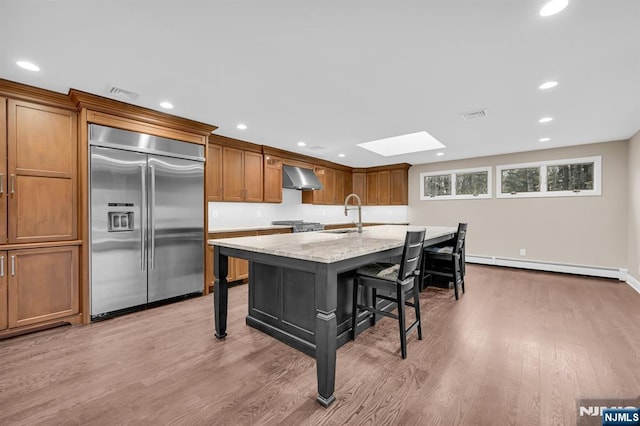 kitchen with built in refrigerator, baseboard heating, wood finished floors, under cabinet range hood, and a kitchen bar