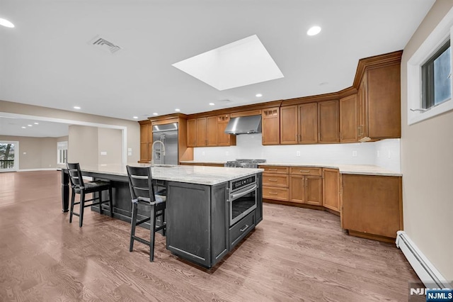 kitchen featuring a skylight, a spacious island, a baseboard radiator, appliances with stainless steel finishes, and ventilation hood