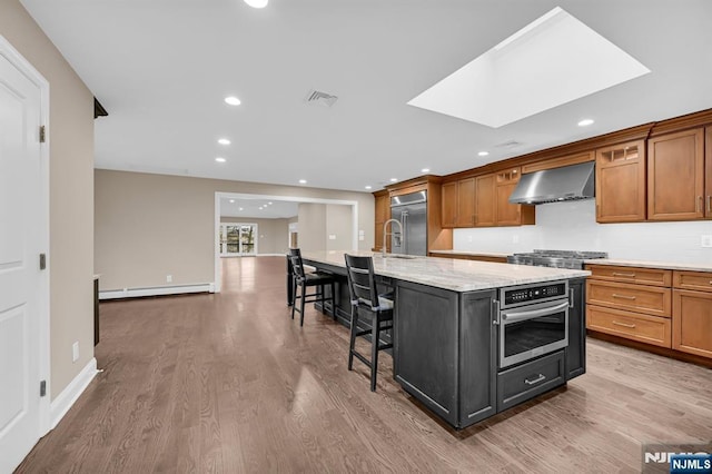 kitchen with a baseboard heating unit, wood finished floors, appliances with stainless steel finishes, wall chimney exhaust hood, and a kitchen bar