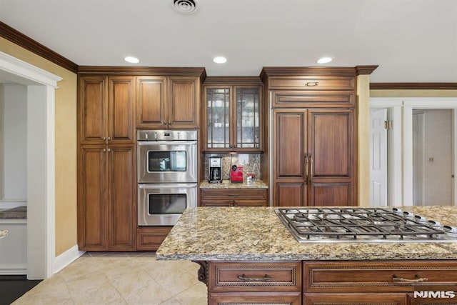 kitchen featuring light stone counters, visible vents, appliances with stainless steel finishes, ornamental molding, and glass insert cabinets