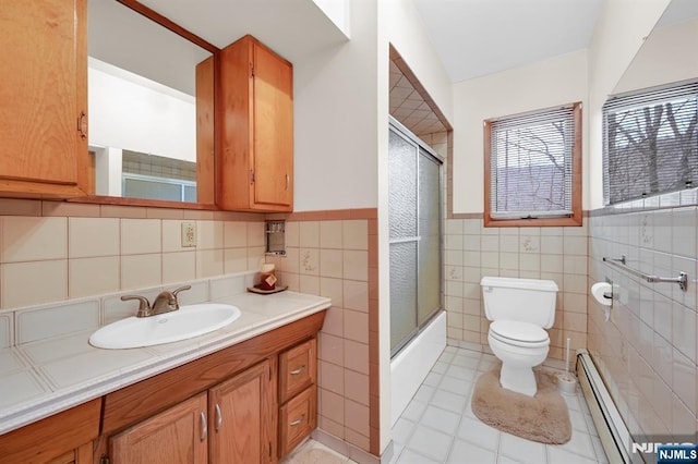 bathroom featuring toilet, shower / bath combination with glass door, vanity, baseboard heating, and wainscoting