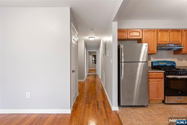 kitchen featuring under cabinet range hood, baseboards, appliances with stainless steel finishes, tasteful backsplash, and brown cabinetry