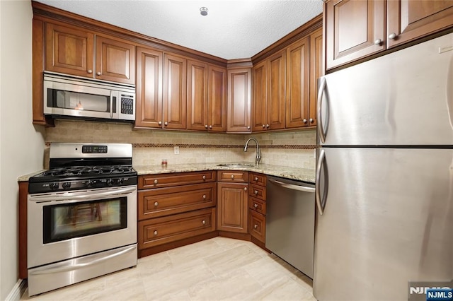 kitchen featuring stainless steel appliances, a sink, brown cabinets, light stone countertops, and tasteful backsplash