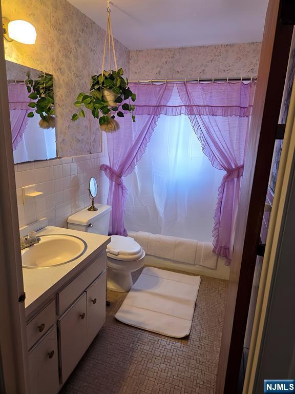 bathroom with toilet, vanity, tile walls, shower / bath combo with shower curtain, and wallpapered walls