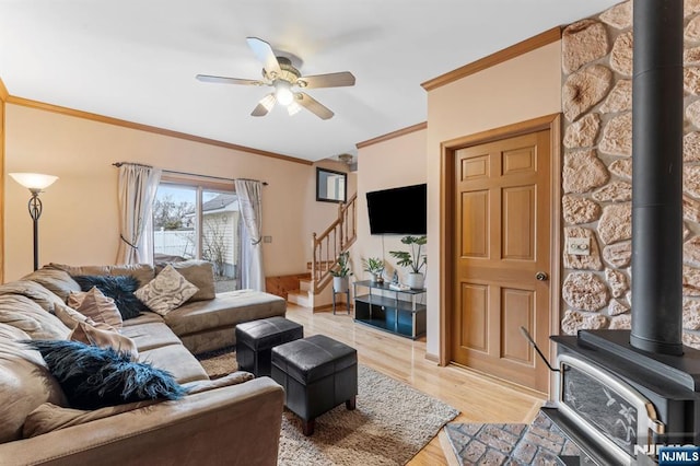 living room with light wood finished floors, ceiling fan, a wood stove, stairs, and crown molding