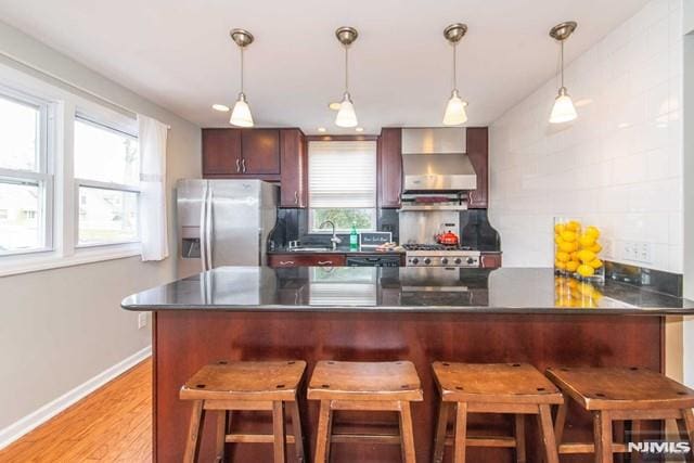 kitchen with dark countertops, decorative backsplash, light wood-style flooring, stainless steel fridge, and wall chimney exhaust hood