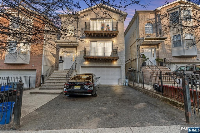 view of property with driveway, a balcony, fence, a garage, and brick siding