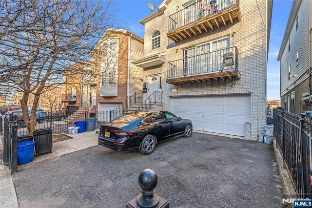 view of property with driveway, a balcony, fence, an attached garage, and brick siding