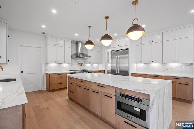 kitchen featuring light wood finished floors, a sink, a large island, built in appliances, and wall chimney exhaust hood