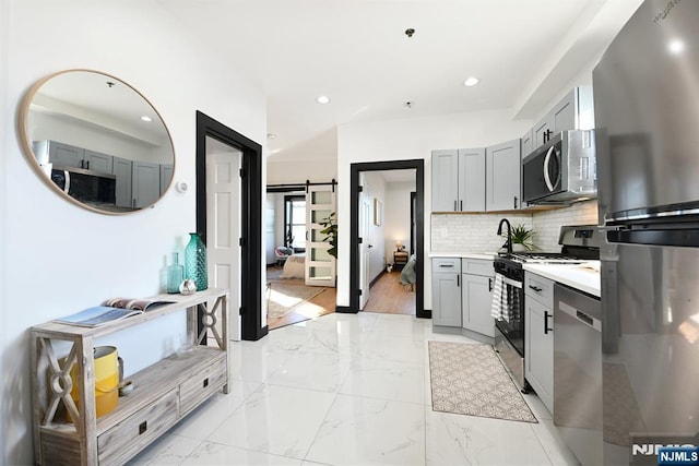 kitchen featuring gray cabinets, decorative backsplash, appliances with stainless steel finishes, a barn door, and marble finish floor